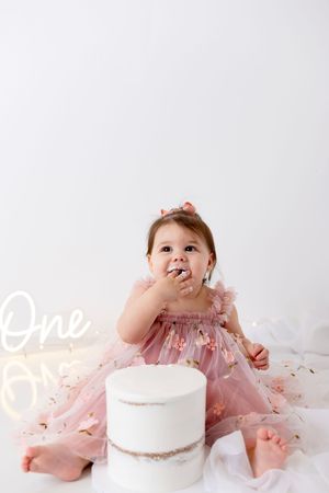 Baby in pink dress sitting on white backdrop with white cake during first birthday cake smash photo session.