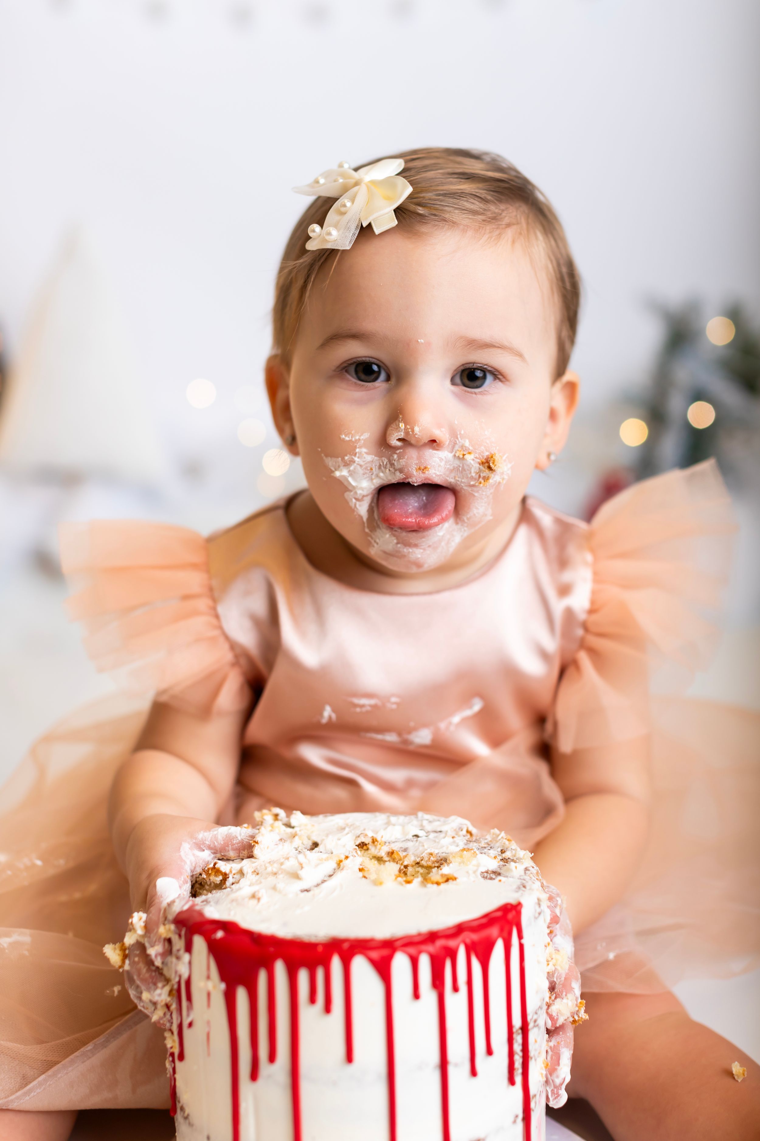 Baby in peach tulle dress smiling with cake frosting on face near drip cake.