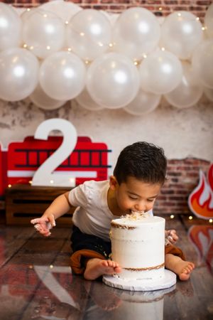 Child leaning over white birthday cake with number 2 and white balloon backdrop.