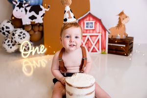 Baby boy in suspenders celebrates first birthday with cake smash, farm theme decor, barn backdrop, and cow print party hat.  NJ photography