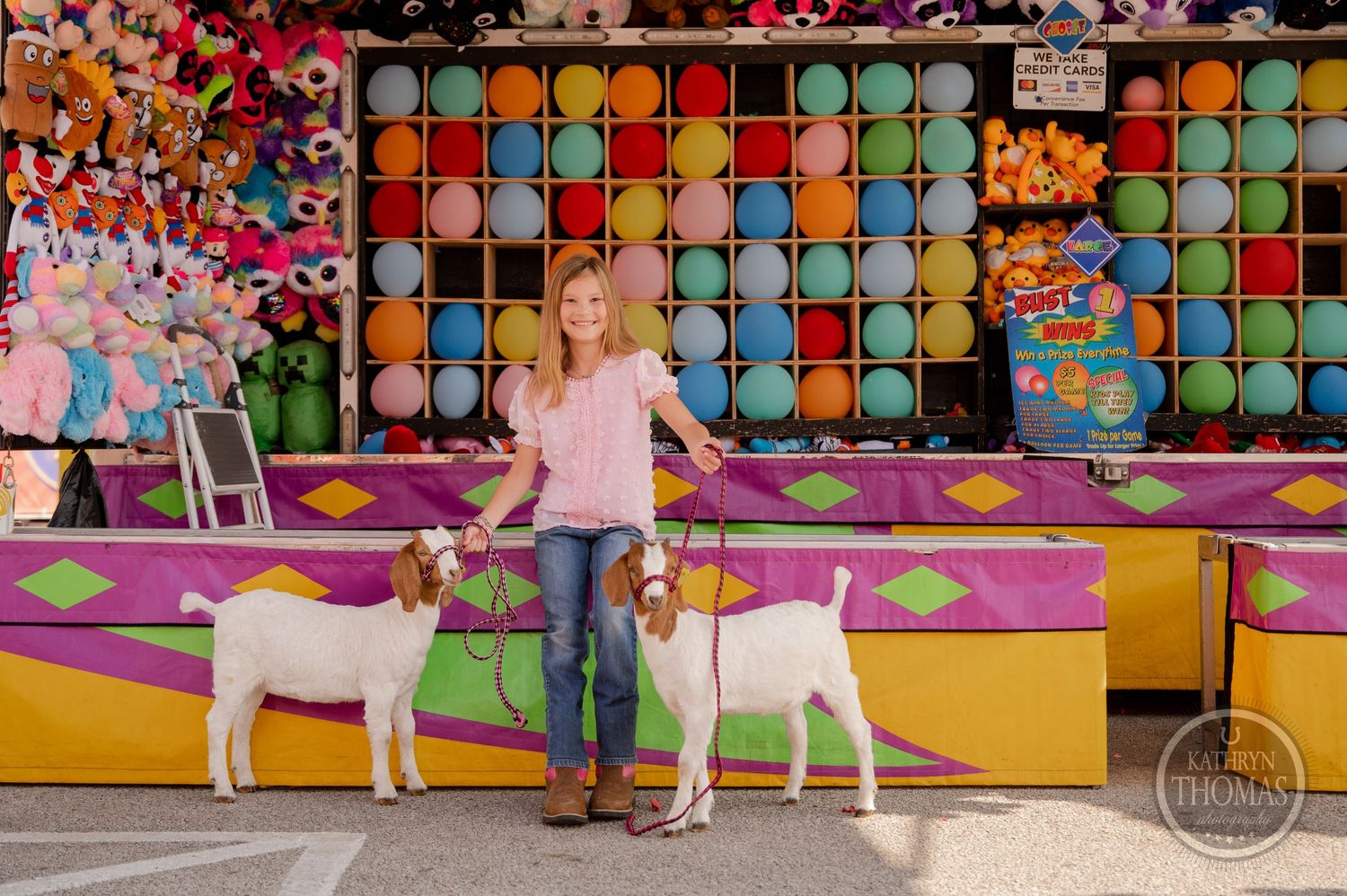 A person stands between two white goats in front of a colorful carnival game booth with balloon targets.