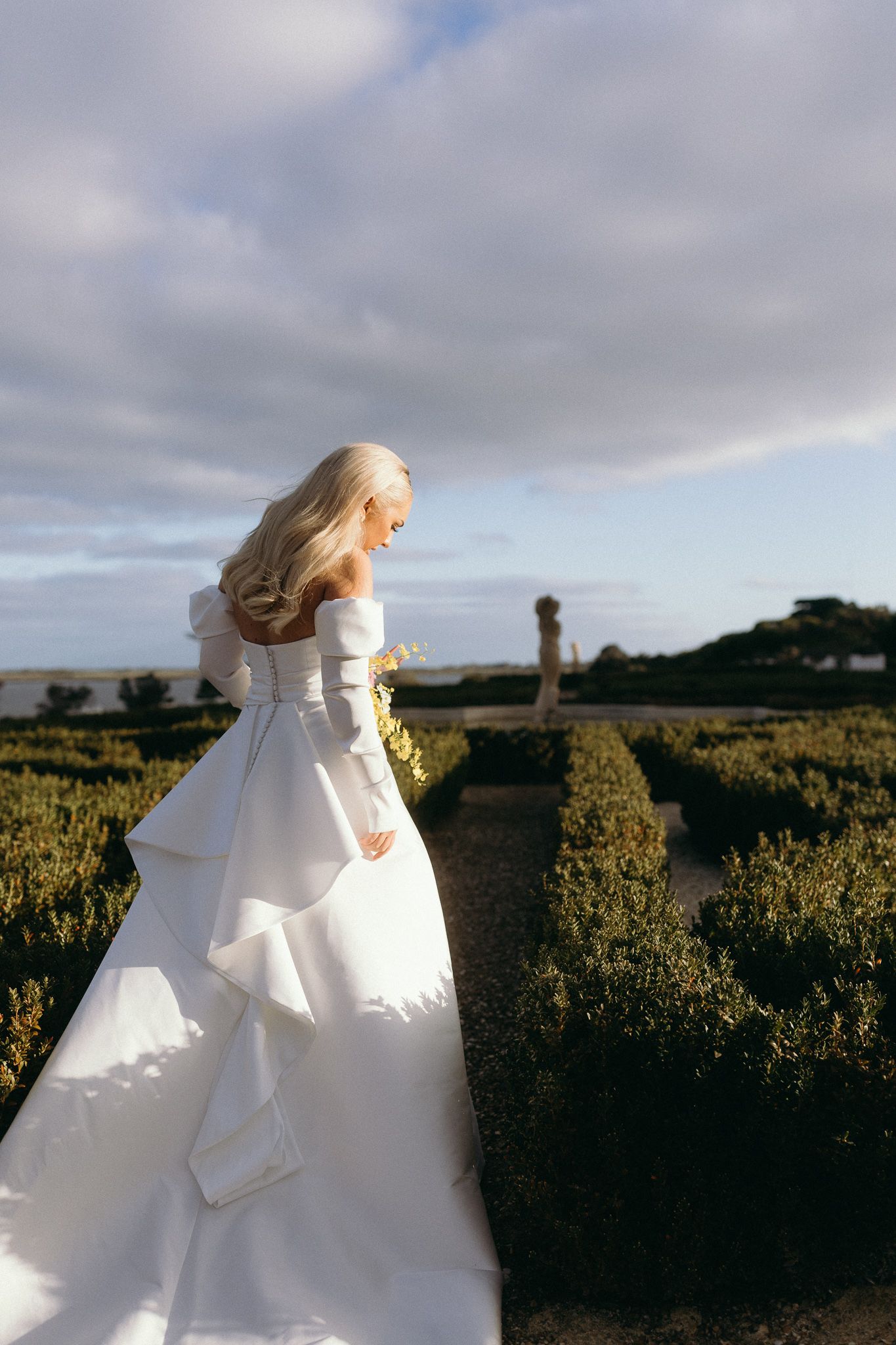 Bride in white satin dress in full sun in hedged garden looking down walking away