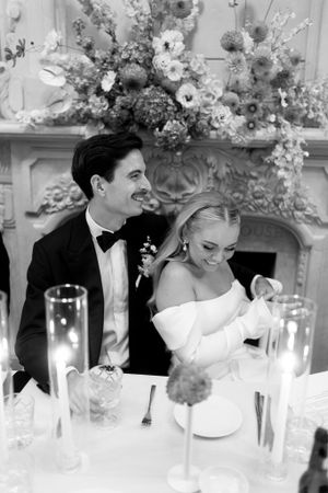 Bride and groom sitting in reception listening to speeches laughing with florals behind them in black and white
