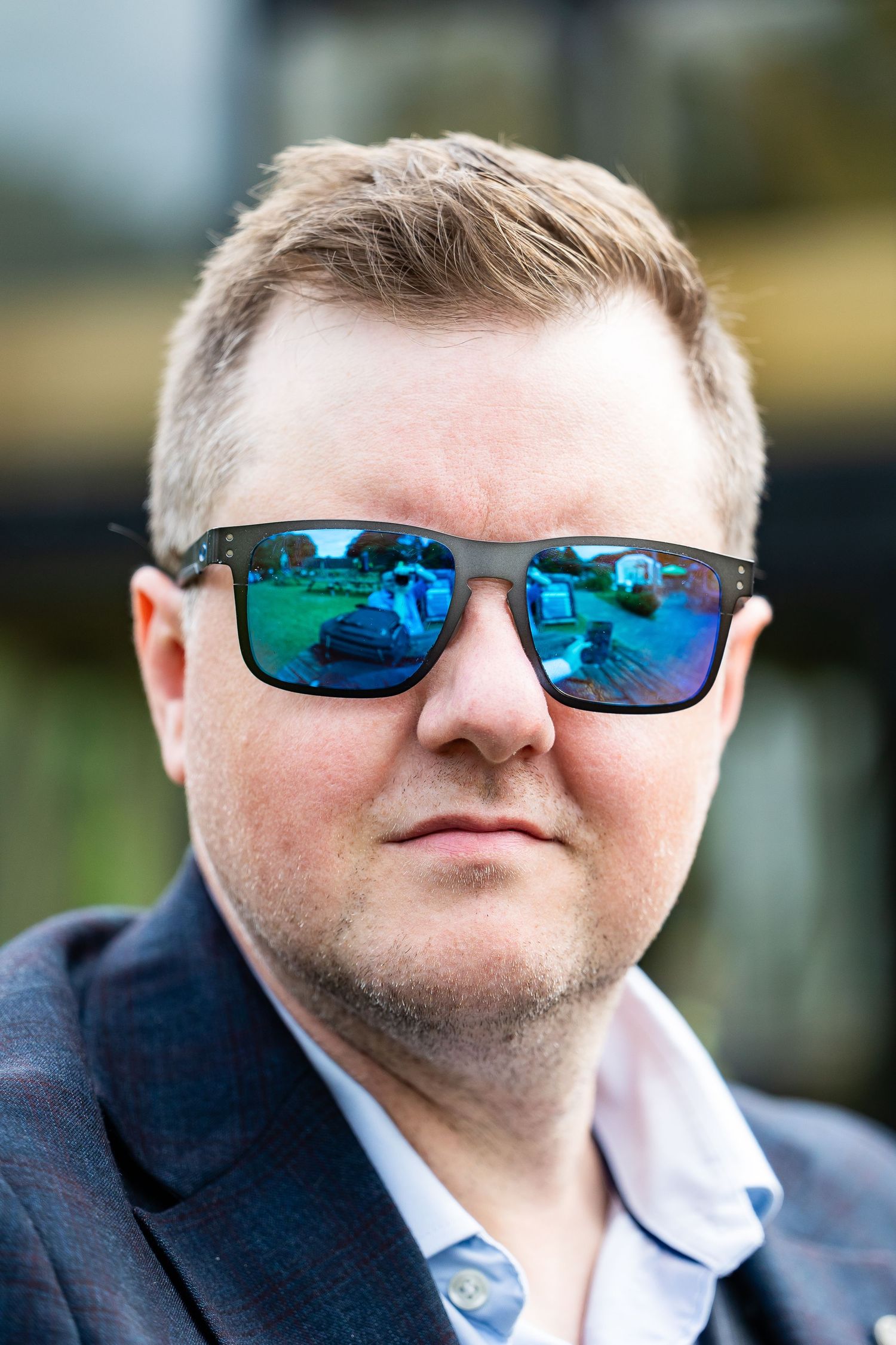 Close-up portrait shot of someone wearing stylish mirrored sunglasses with blue-green lenses and a dark blazer.