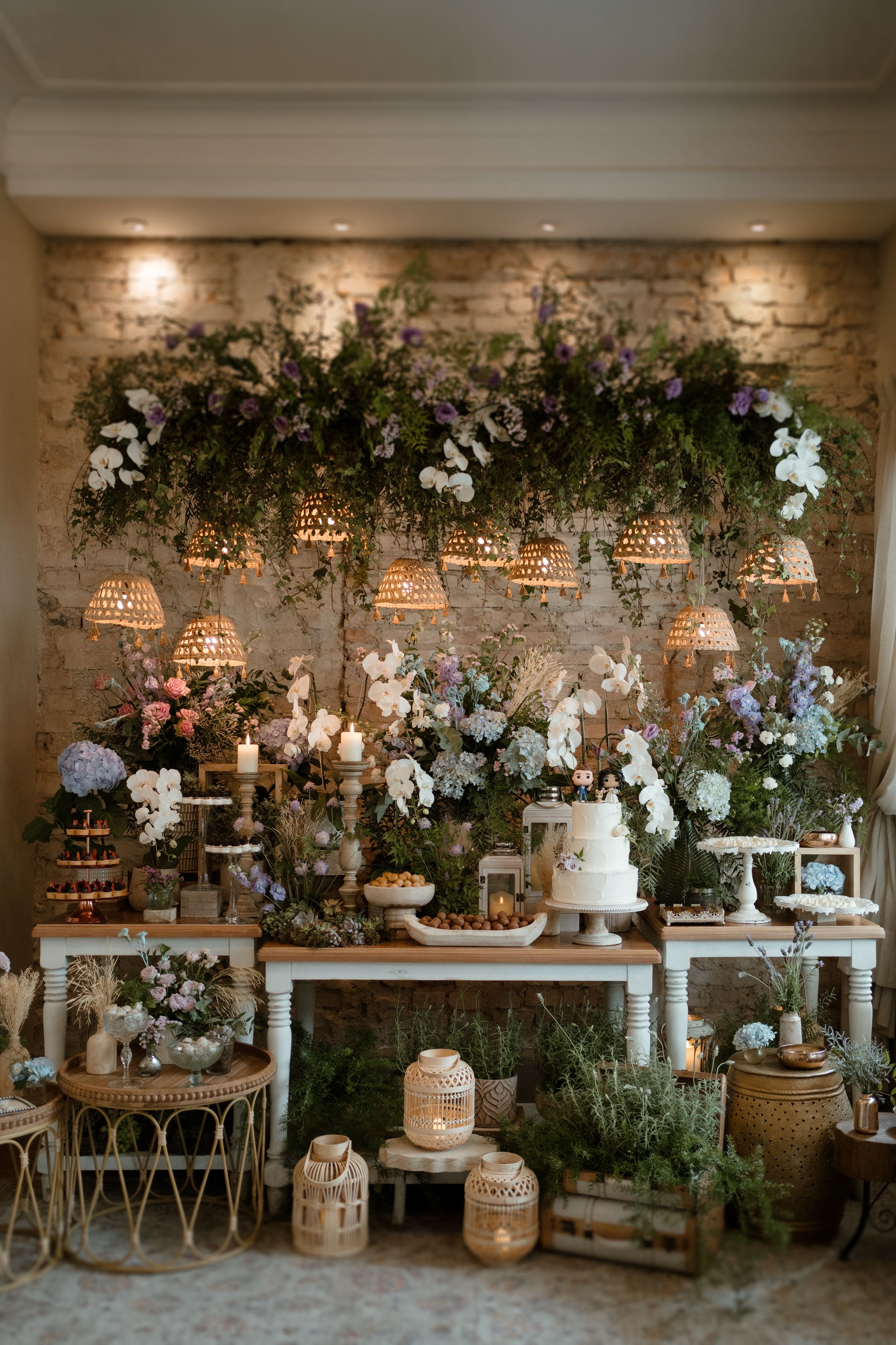 Elegant wedding dessert table with white flowers, greenery and rustic decor against a brick wall.