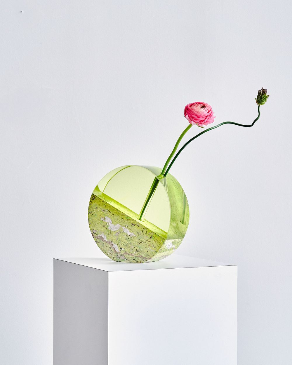 Green glass sculpture with pink flower stem balanced on white display platform.