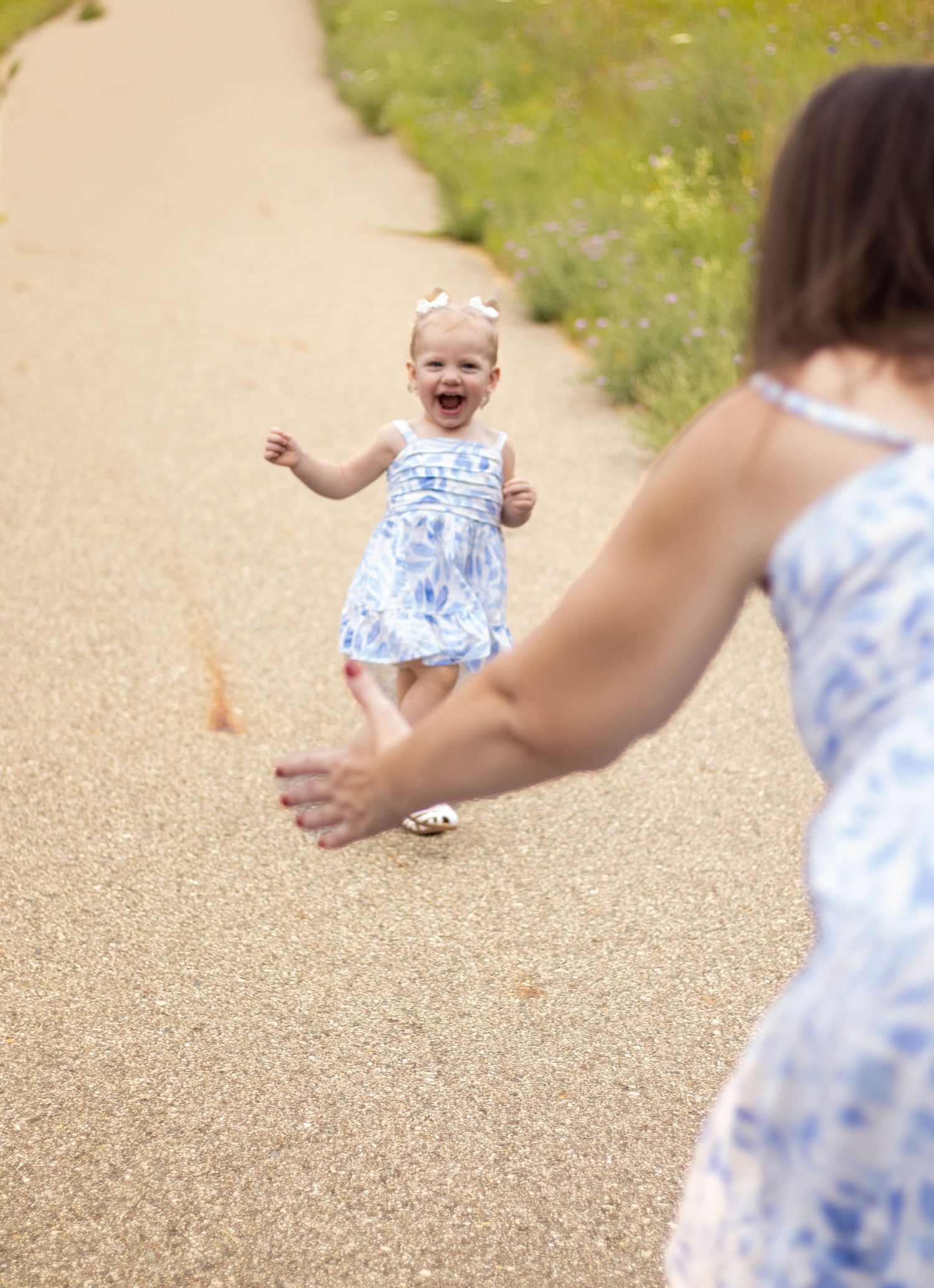 A joyful child in a floral dress runs along a gravel path on a sunny day while playing with others.