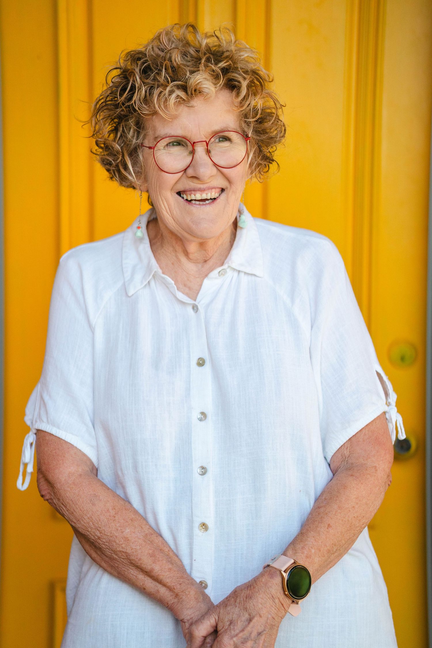 A warm portrait against a yellow door features round glasses and a crisp white button-down shirt.