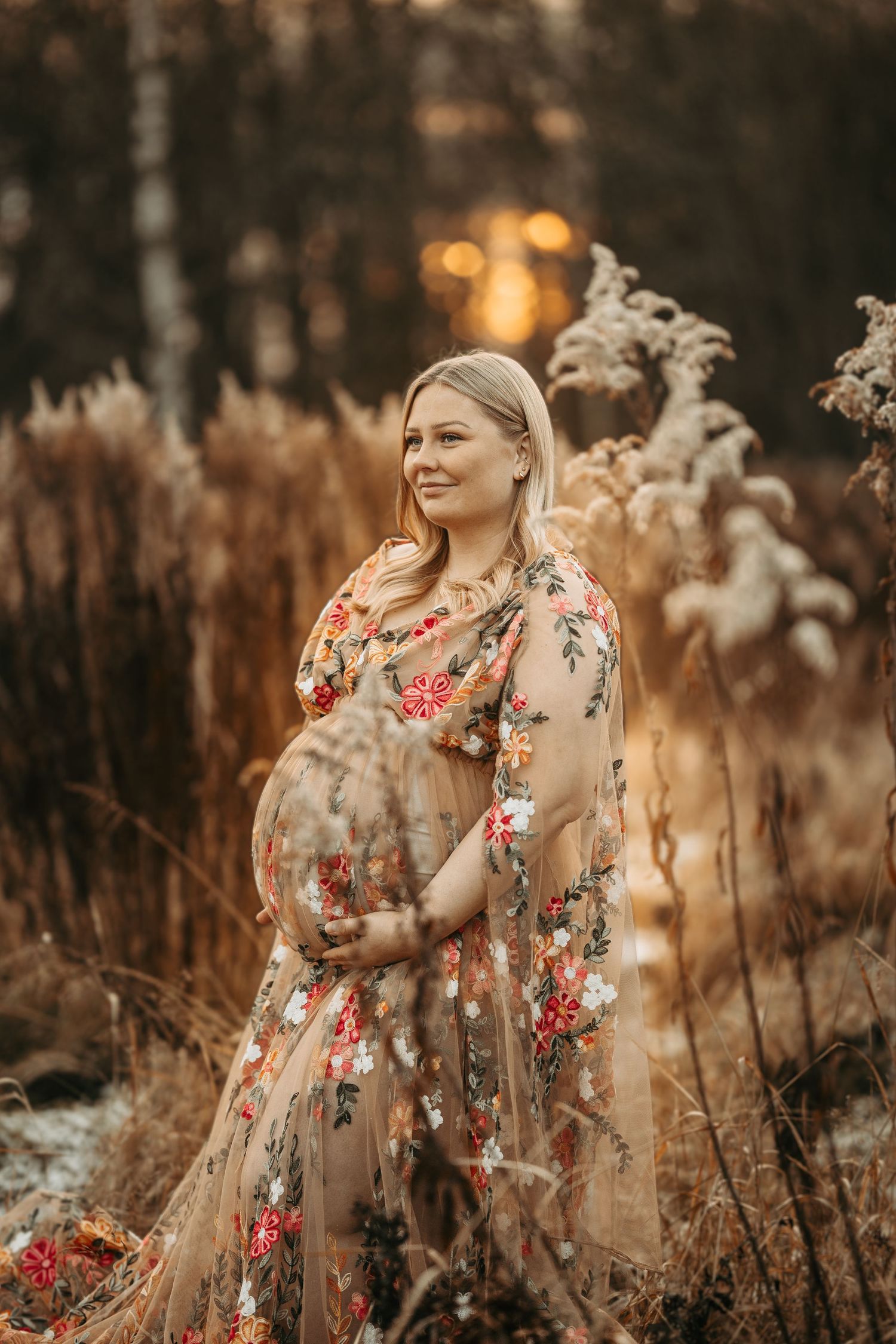 A person in a flowing floral embroidered gown poses on a beach at sunset with warm golden tones.