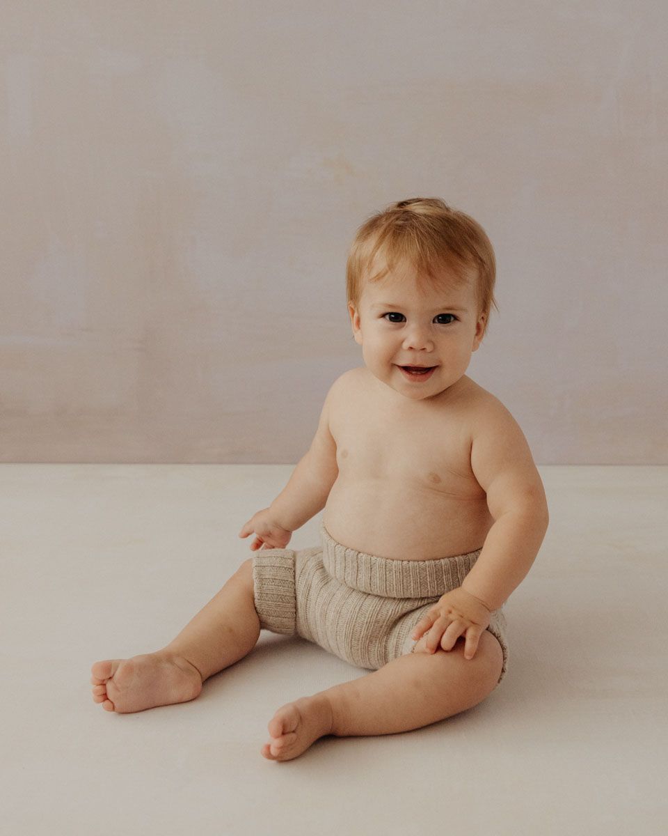 Baby in knitted diaper cover poses on cream backdrop in minimalist photography series.