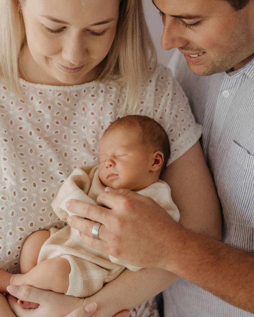 Parents in neutral colors holding newborn baby during intimate indoor photo session.