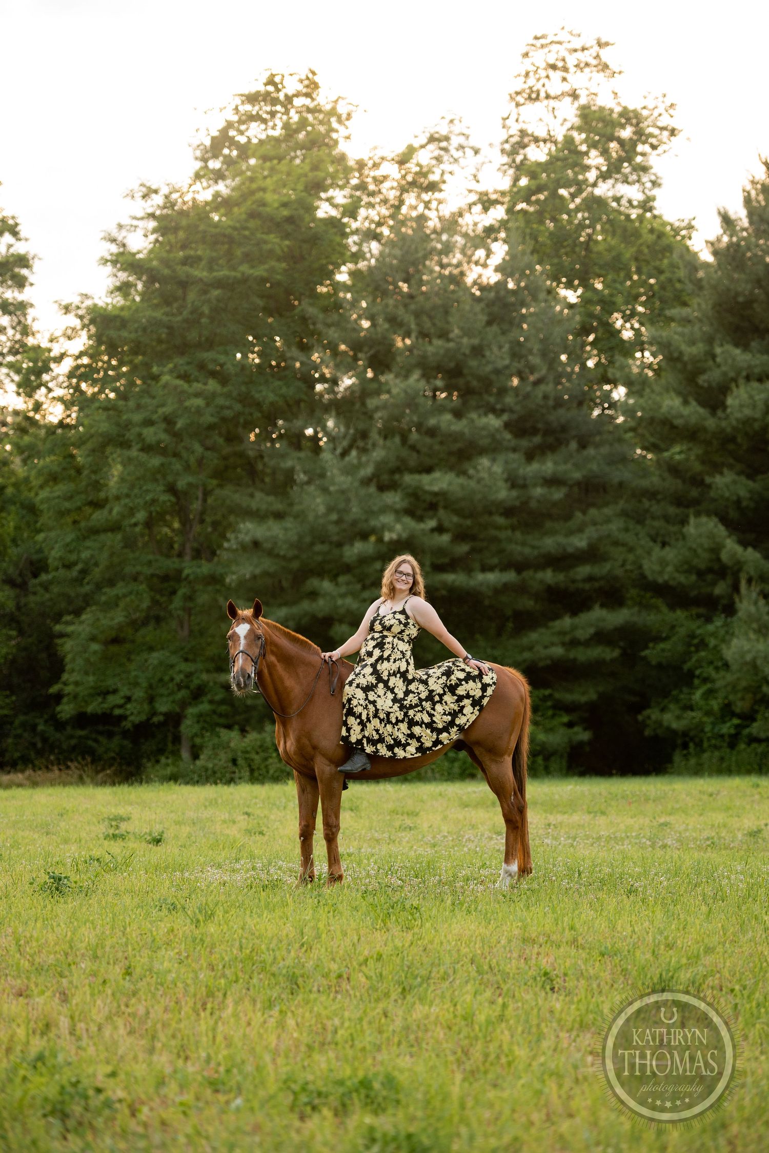 A chestnut horse stands in a grassy field at sunset with a rider in a floral dress against a backdrop of evergreen trees.