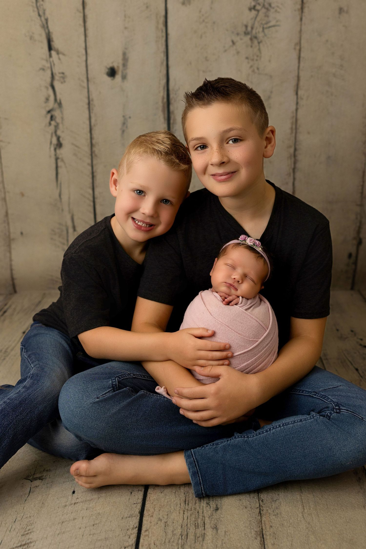 Three siblings in matching black shirts and jeans pose lovingly together with a newborn baby on a rustic wood floor.