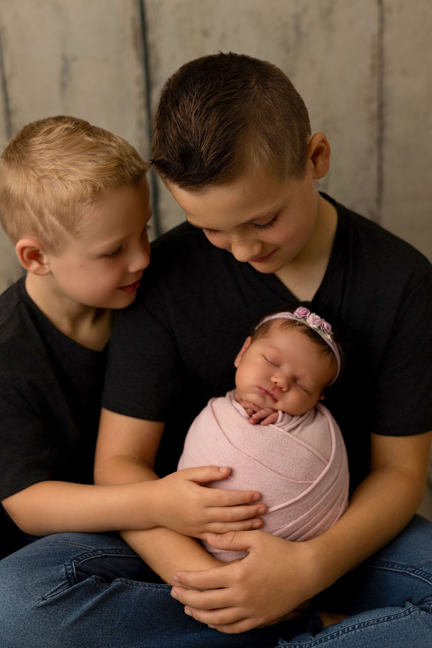 A loving moment captured in black and white as siblings bond with a swaddled newborn baby in pink.