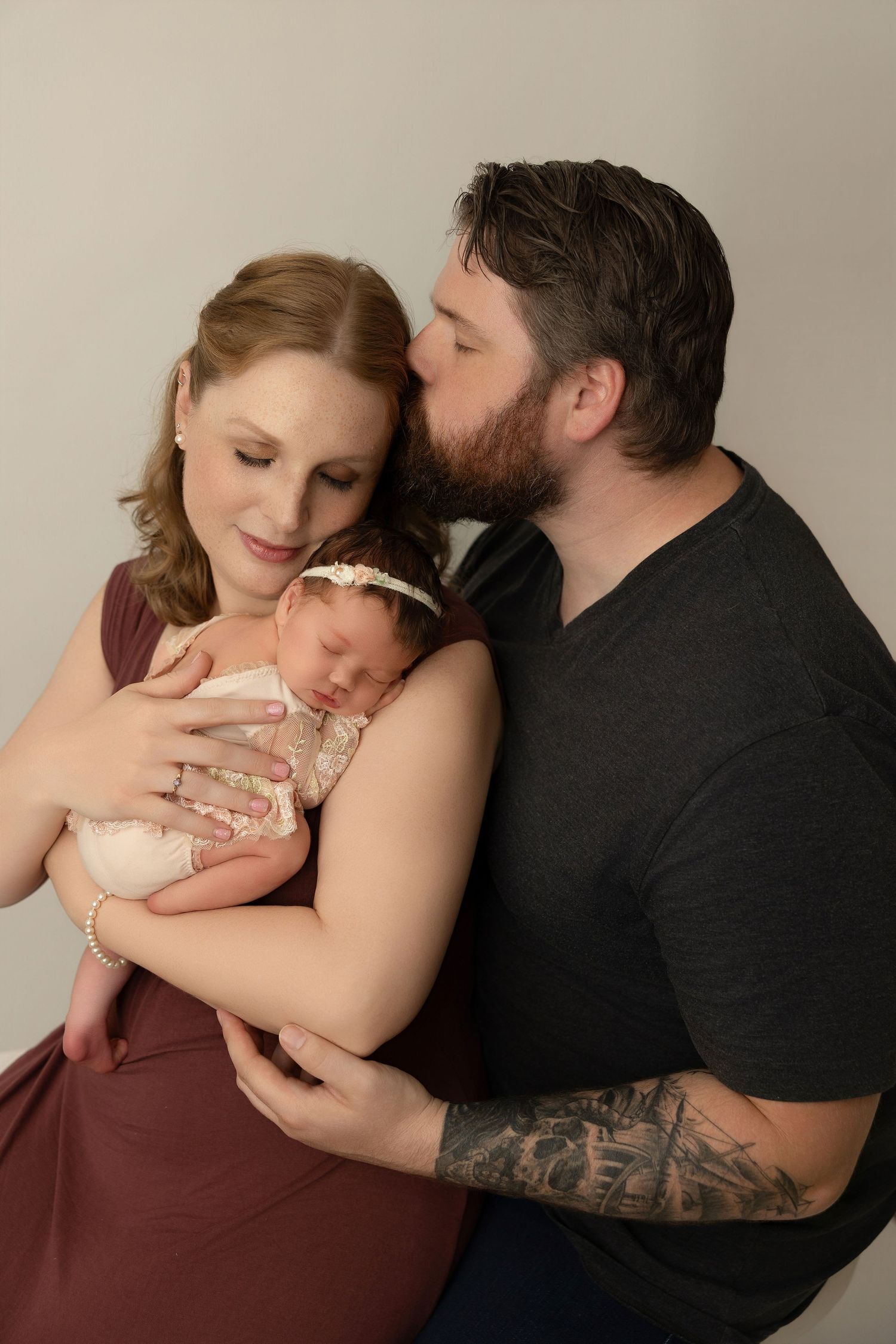 Studio photography series showing couple embracing while holding their newborn baby in burgundy and black outfits.