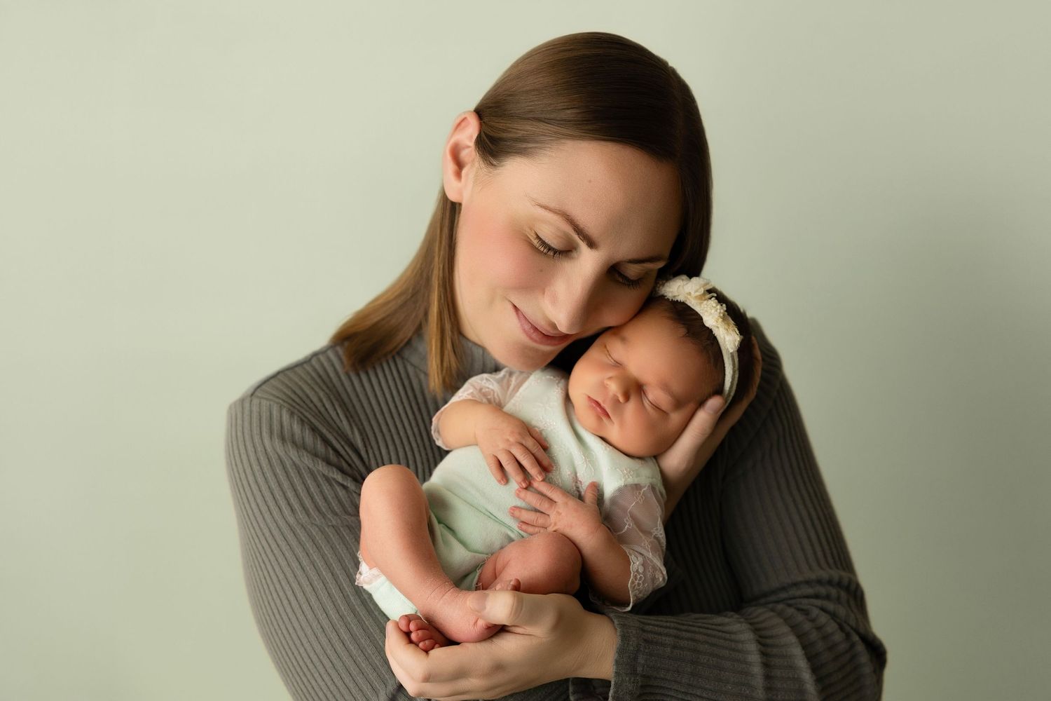 A mother in a gray sweater tenderly holds and snuggles her sleeping newborn baby against her chest.