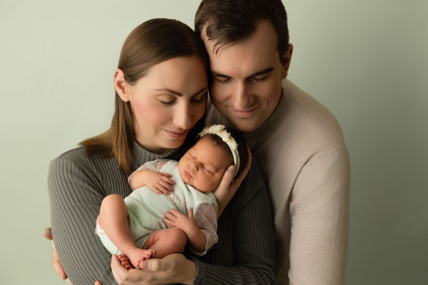 A couple in cozy sweaters tenderly holds their sleeping newborn baby against a neutral background.