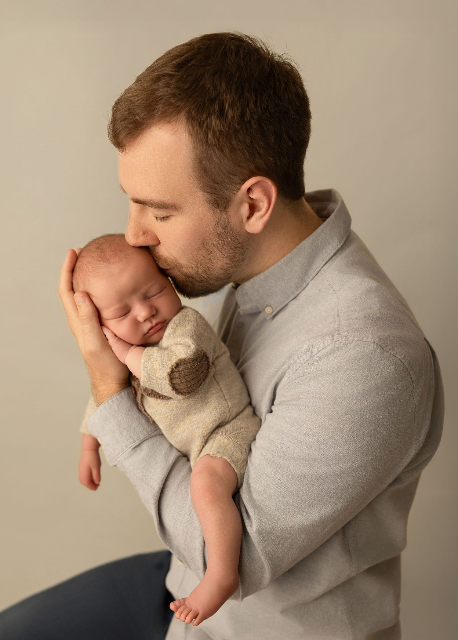 A tender moment between parent and newborn baby wearing beige outfit against neutral background.