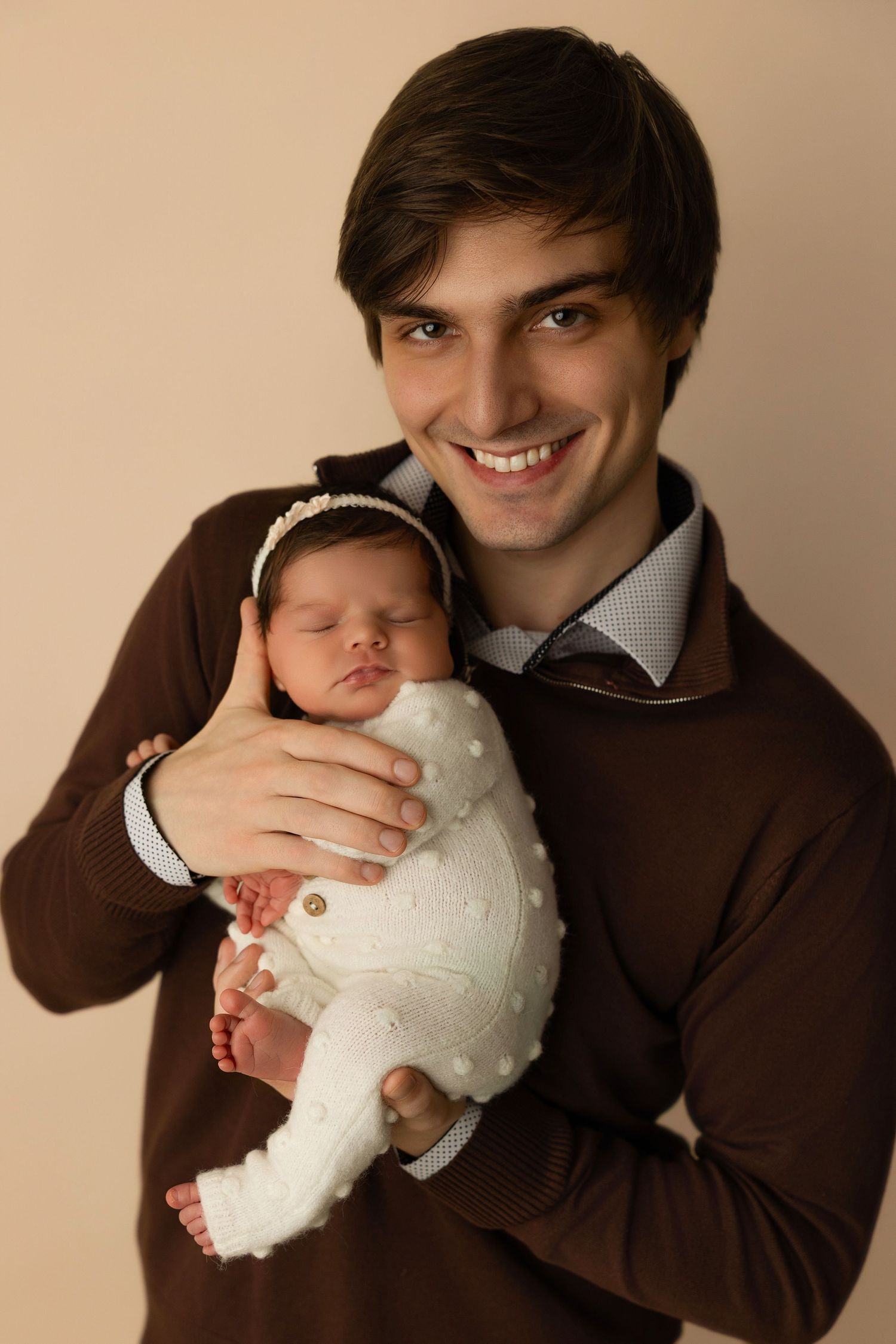 A sweet newborn baby wearing a white dress is cradled in someone's arms wearing a brown sweater.