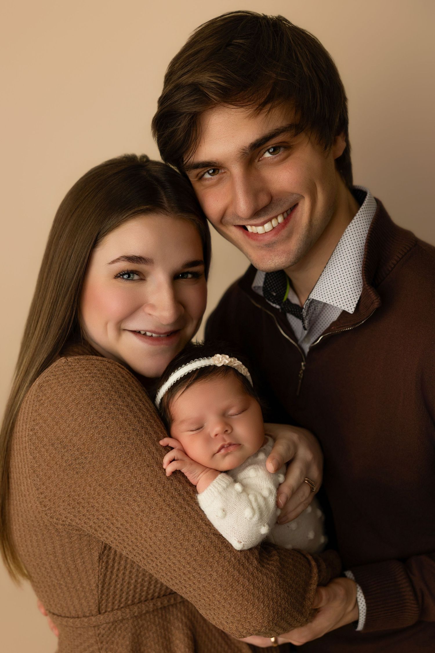 Professional studio portrait of a couple in brown sweaters holding their sleeping newborn baby wearing white and a headband.