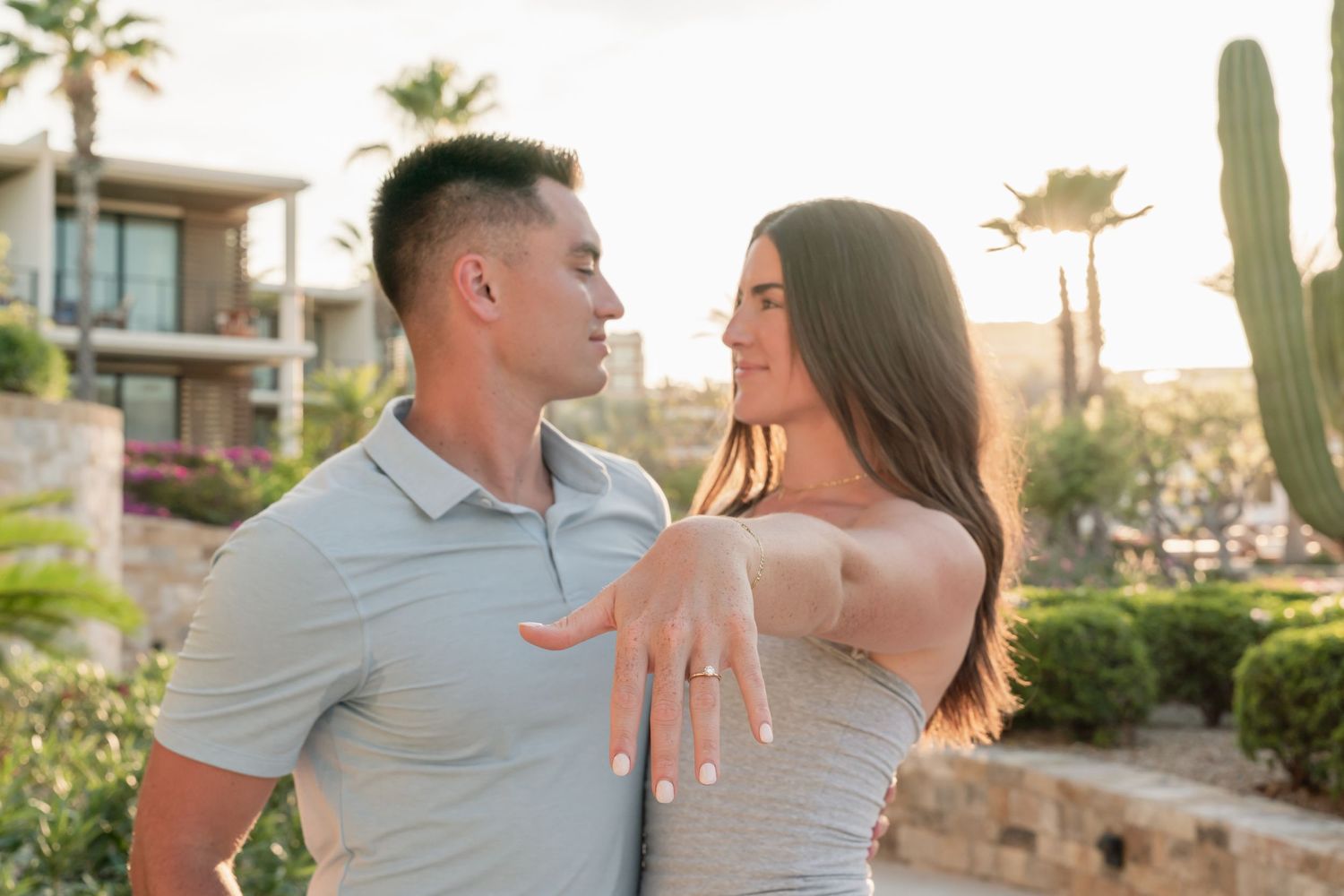 Couple shows off engagement ring at sunset with desert landscape and palm trees in background.