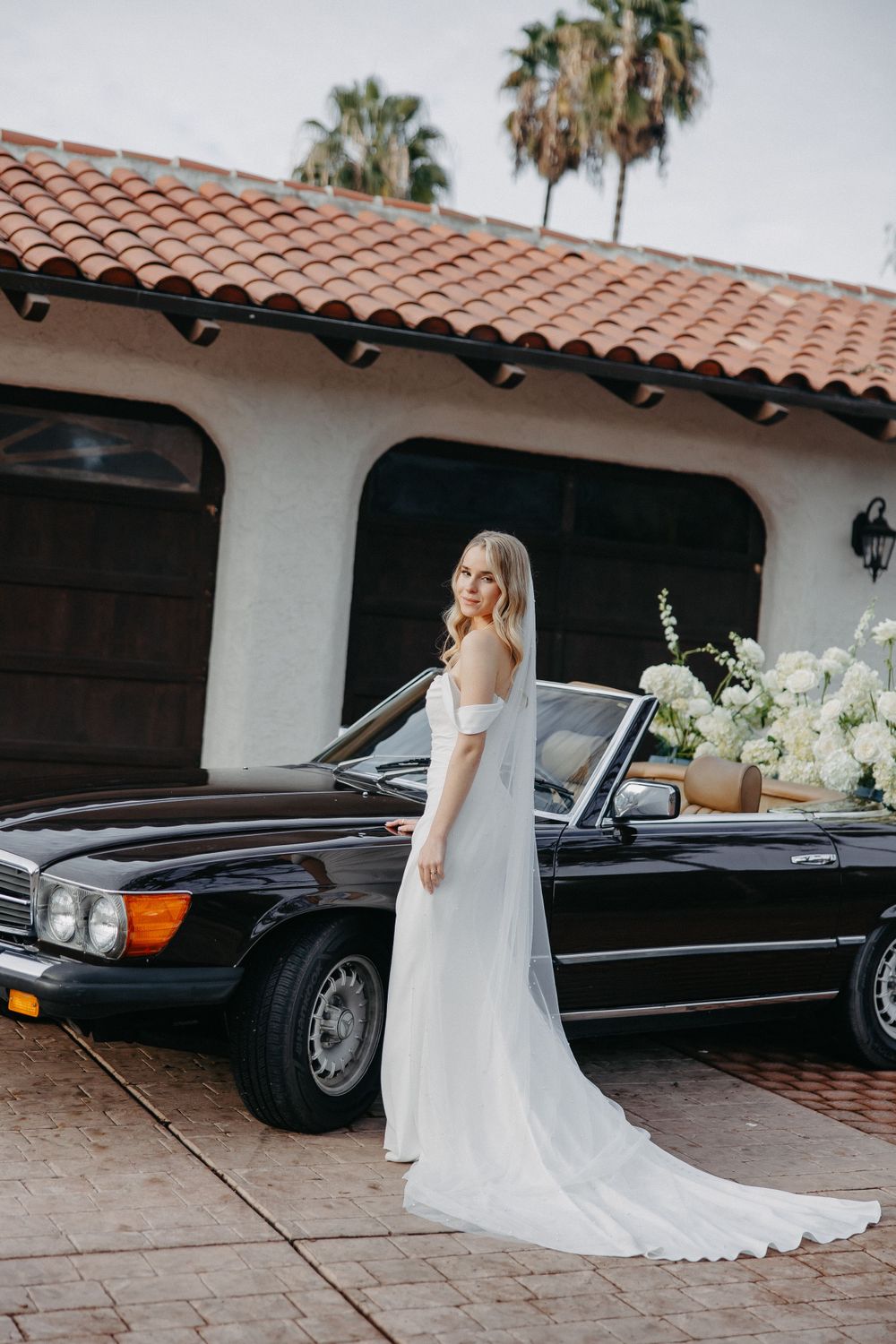 Bride in sleek white gown poses beside vintage black Mercedes convertible.