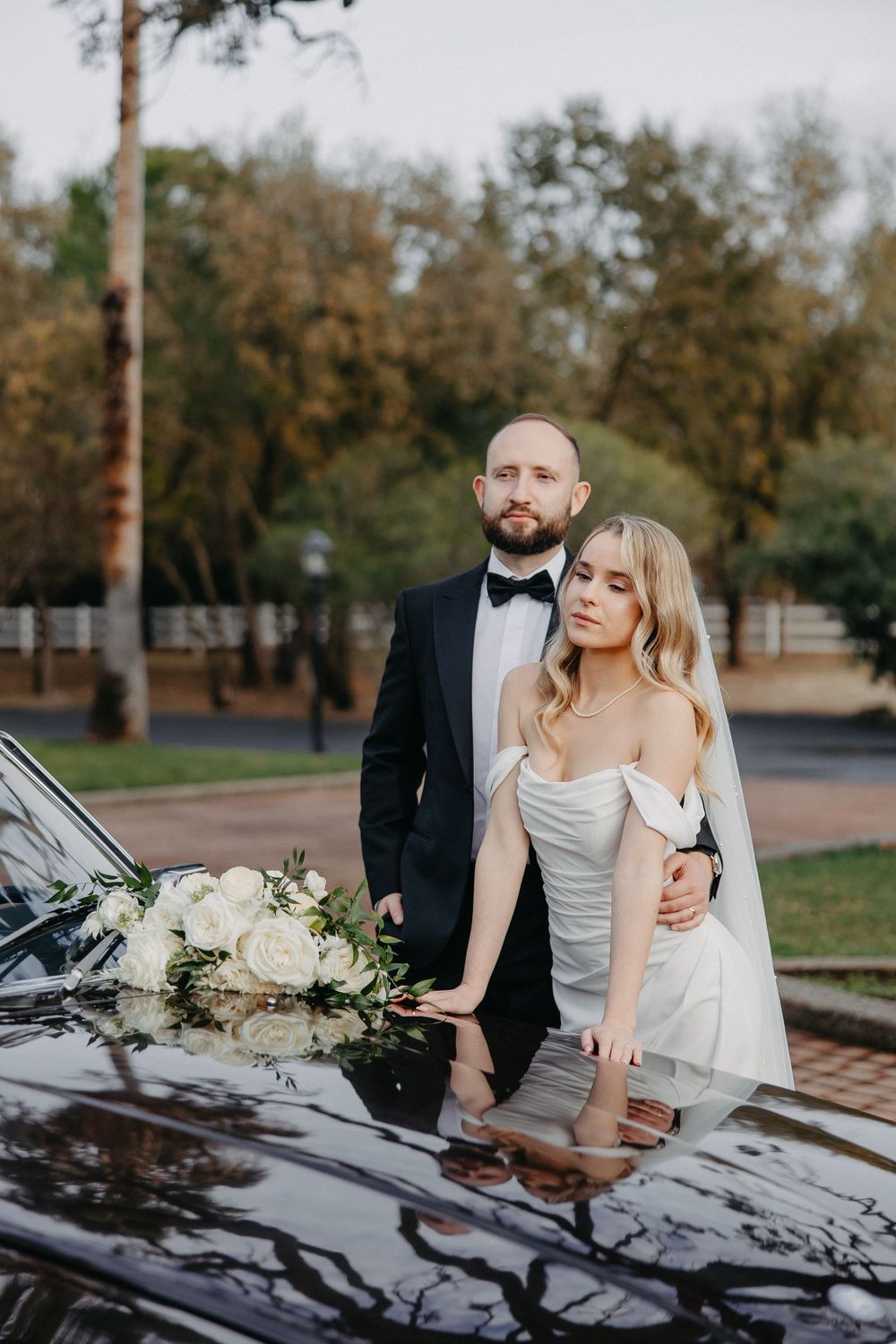 Wedding couple leaning on classic black car with white floral bouquet.