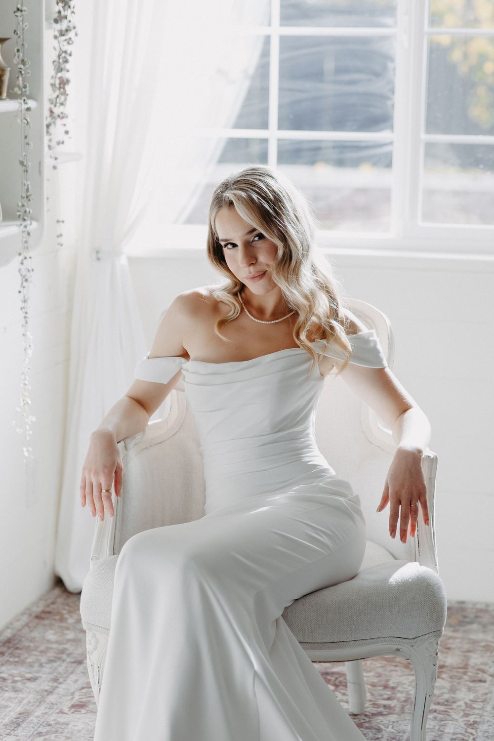 Bride in elegant off-shoulder white dress poses in bright window-lit room.