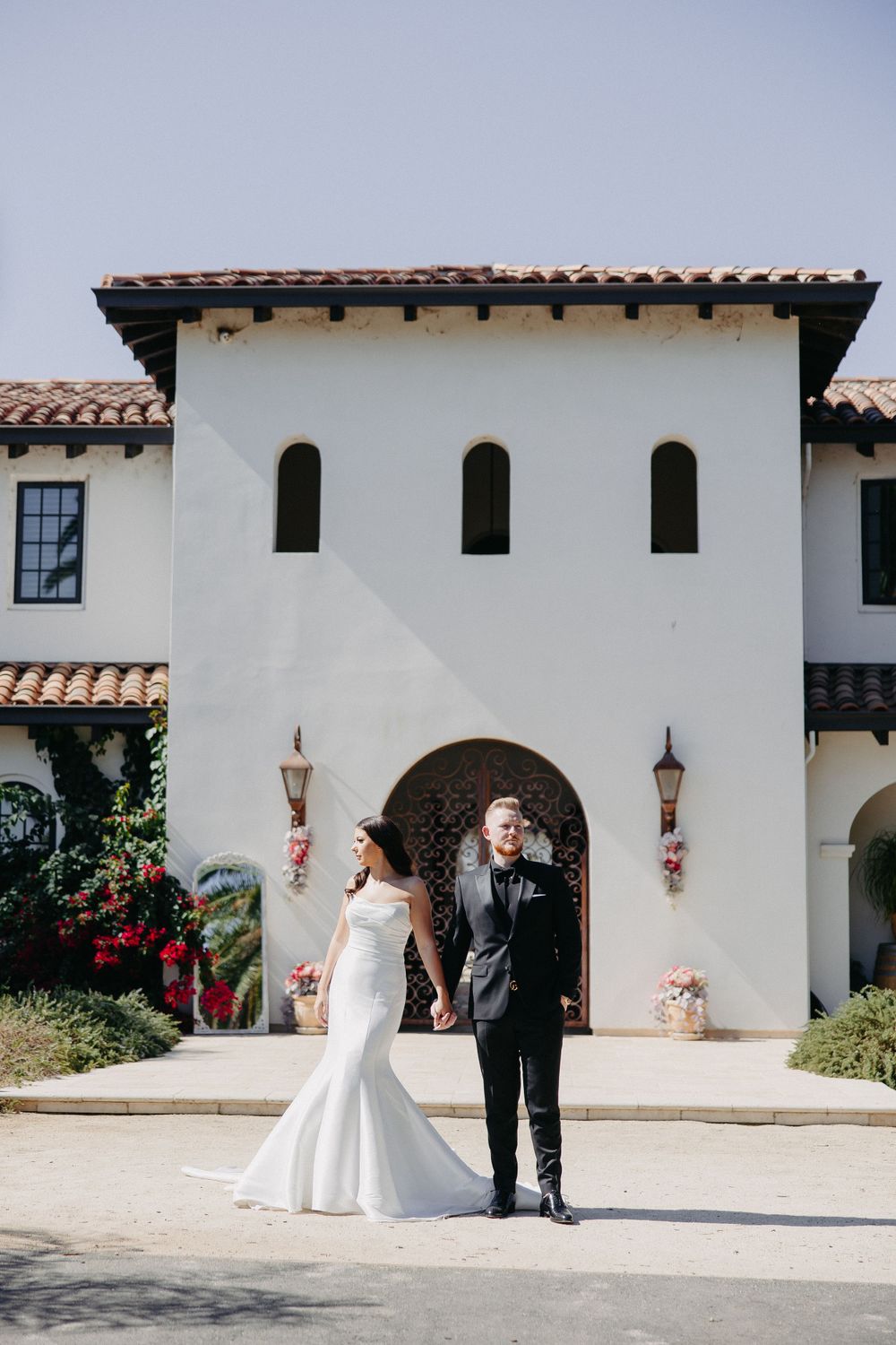 A newlywed couple stands in front of a Spanish-style villa with arched doorways and terracotta roof.