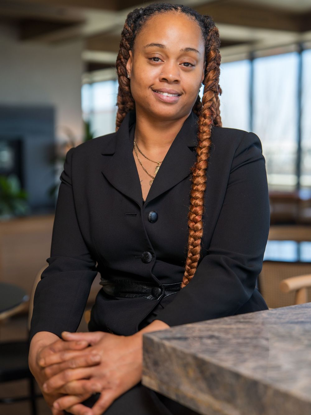 Professional wearing a black blazer sits at a desk in a bright modern office setting with a warm smile.
