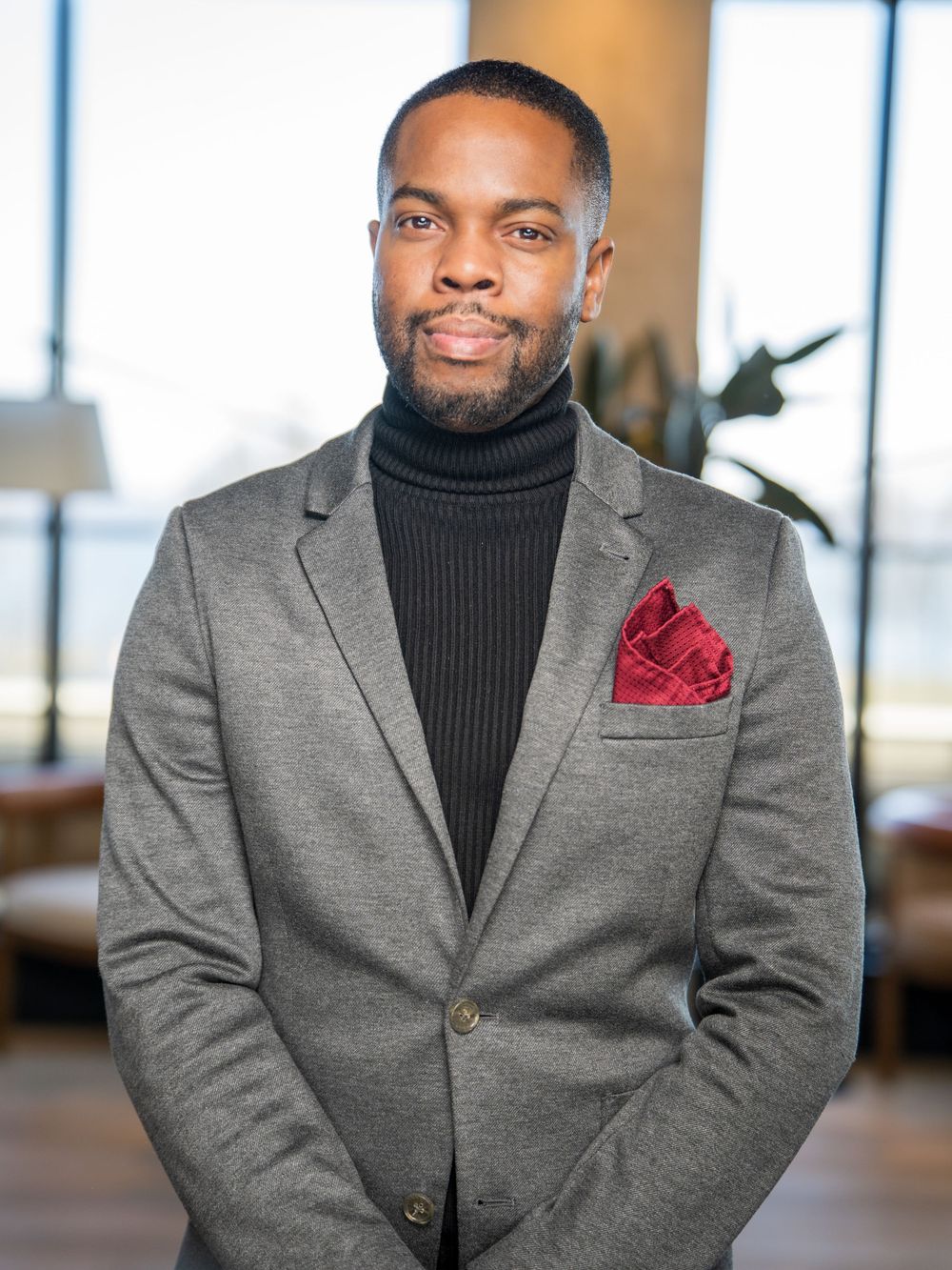 Professional portrait of a person wearing a gray blazer, black turtleneck and red pocket square in an office setting.