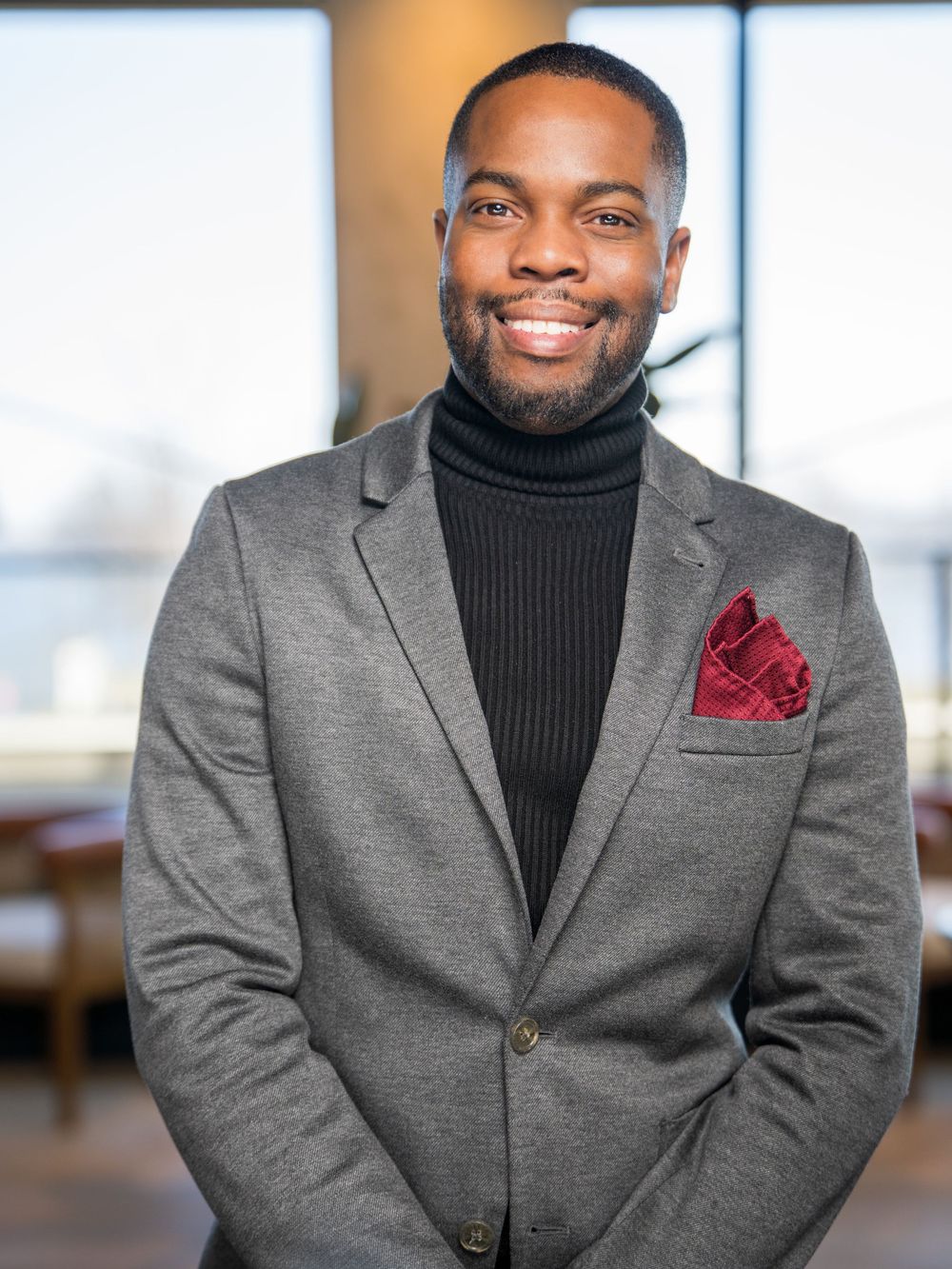 Professional wearing a black turtleneck and gray blazer with burgundy pocket square in an office setting.
