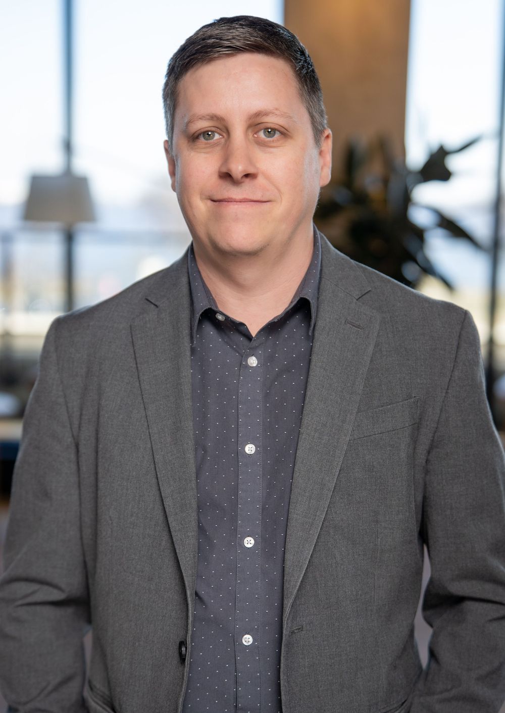 Professional wearing a grey suit jacket and dark polo shirt smiles warmly in an office setting.