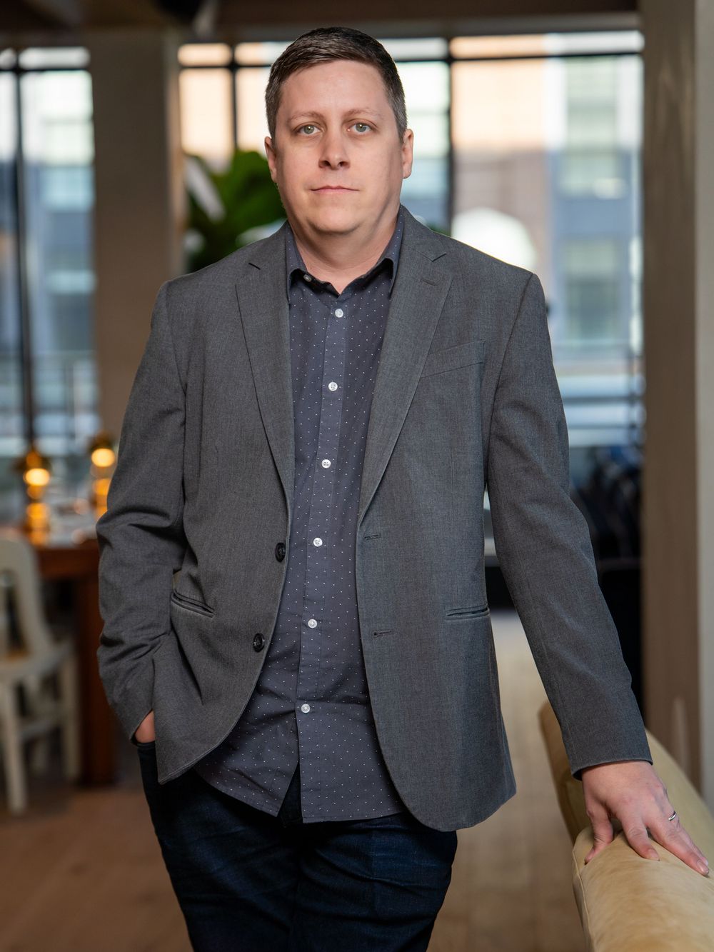Professional wearing a gray suit jacket and navy shirt stands in an office setting with natural lighting.