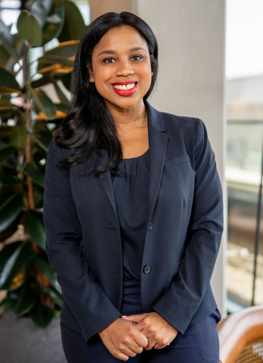 Professional business portrait of someone in a navy blazer smiling warmly in an office setting with plants in background.