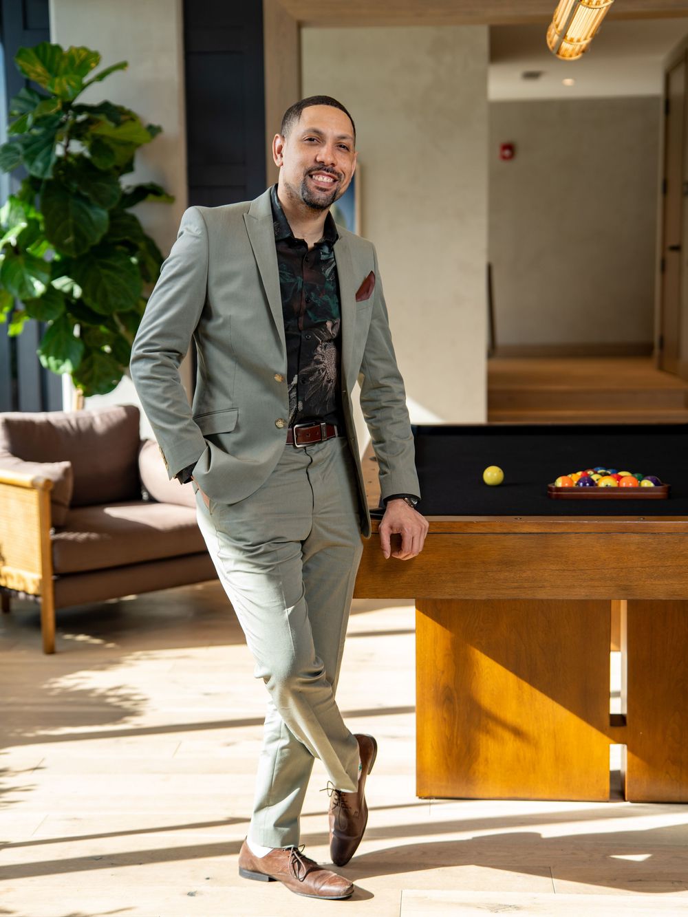 Professional wearing a light gray suit poses playfully near a wooden table in a sunlit indoor space.