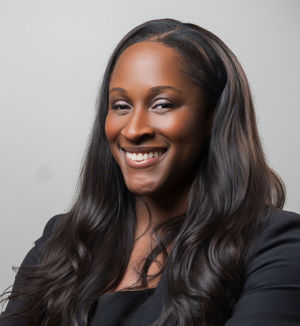A person with long dark hair wearing a black blazer smiles warmly in a professional portrait series.