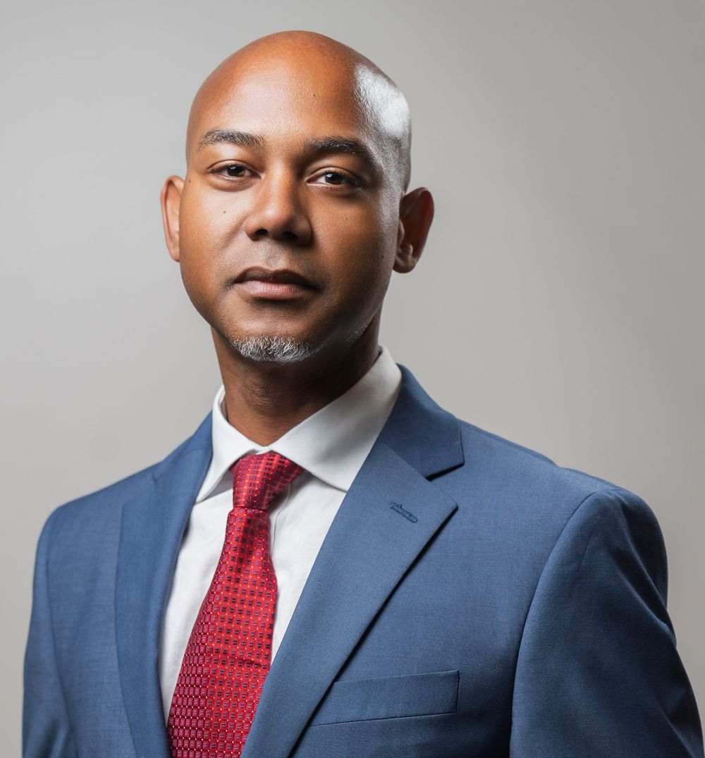 Professional headshot of someone wearing a navy suit and red tie against a light gray background.