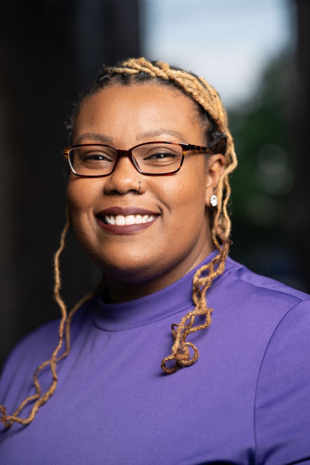 Professional in purple top with blonde braids and glasses smiling warmly at camera.