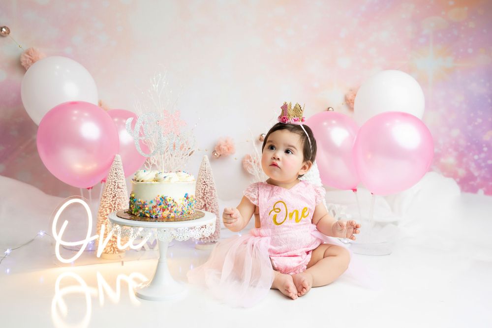 Birthday setup with cake, pink balloons and neon 'One' sign in pastel colors.