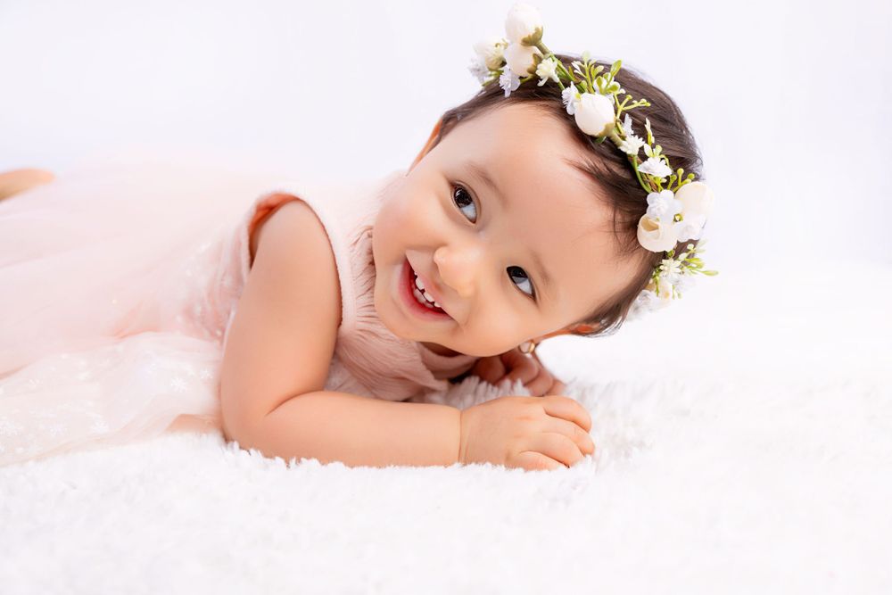 A baby wearing a floral crown and light pink dress lies on white fur bedding with a gentle smile.