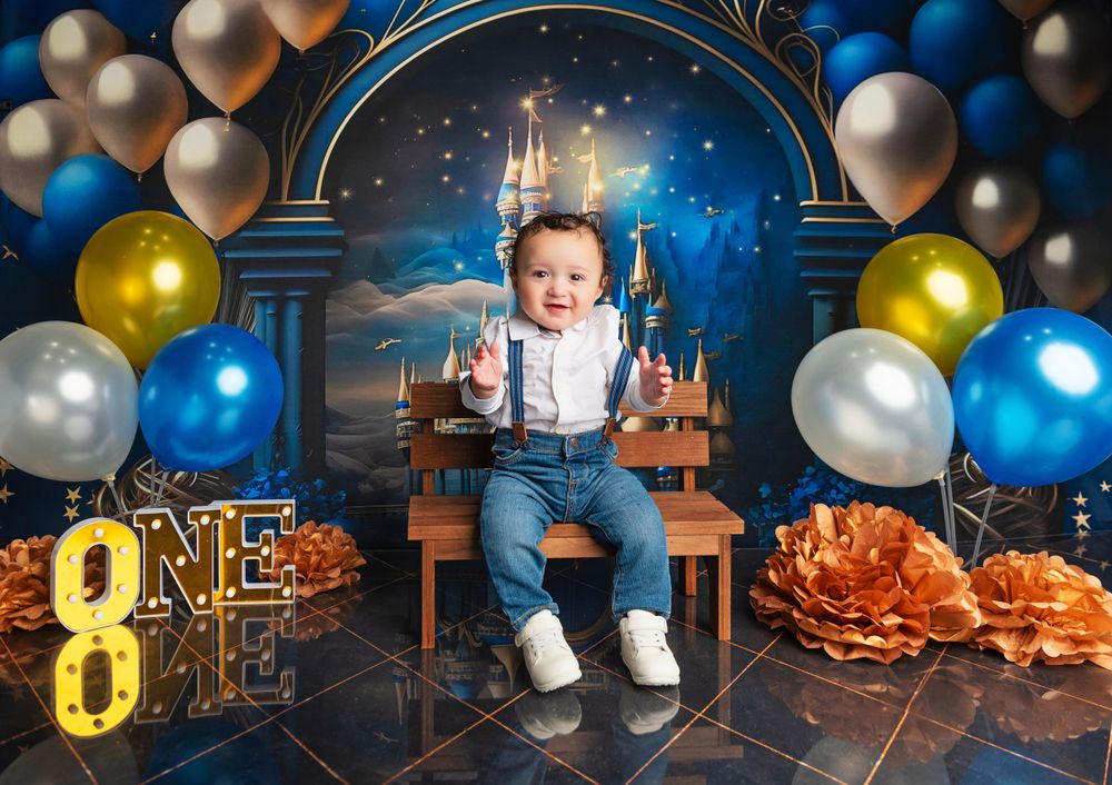 One year old in suspenders smiling on themed birthday set with balloons and castle backdrop.