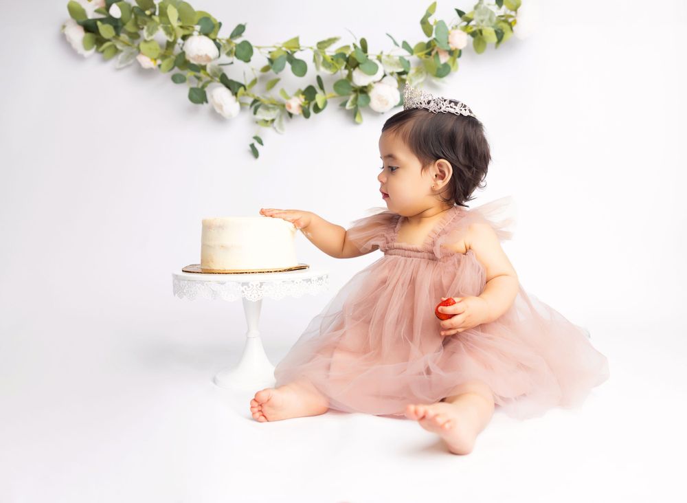 Baby in pink tulle dress reaching for cake against white backdrop with eucalyptus garland.