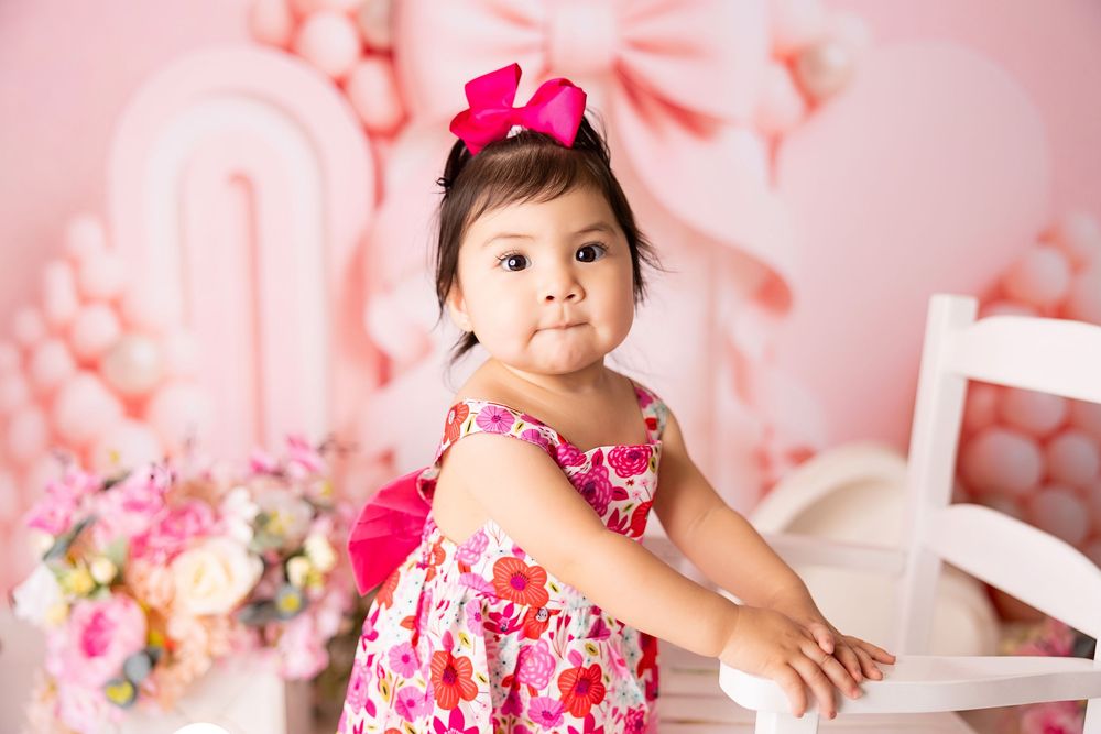 Adorable toddler girl in floral pink dress with bow headband poses in a pink Valentine's Day photo backdrop.