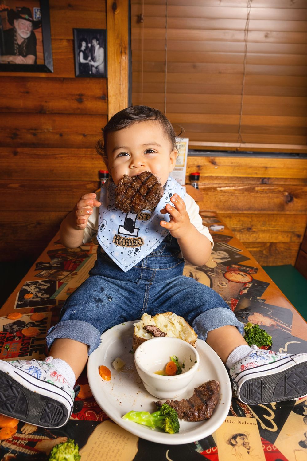 Baby in denim overalls and rodeo bandana holds a steak at a rustic restaurant with a plate of food nearby.