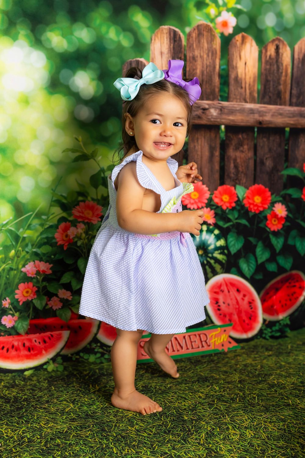 A baby in a purple gingham dress poses with watermelon slices in a garden setting with red flowers and wooden fence.