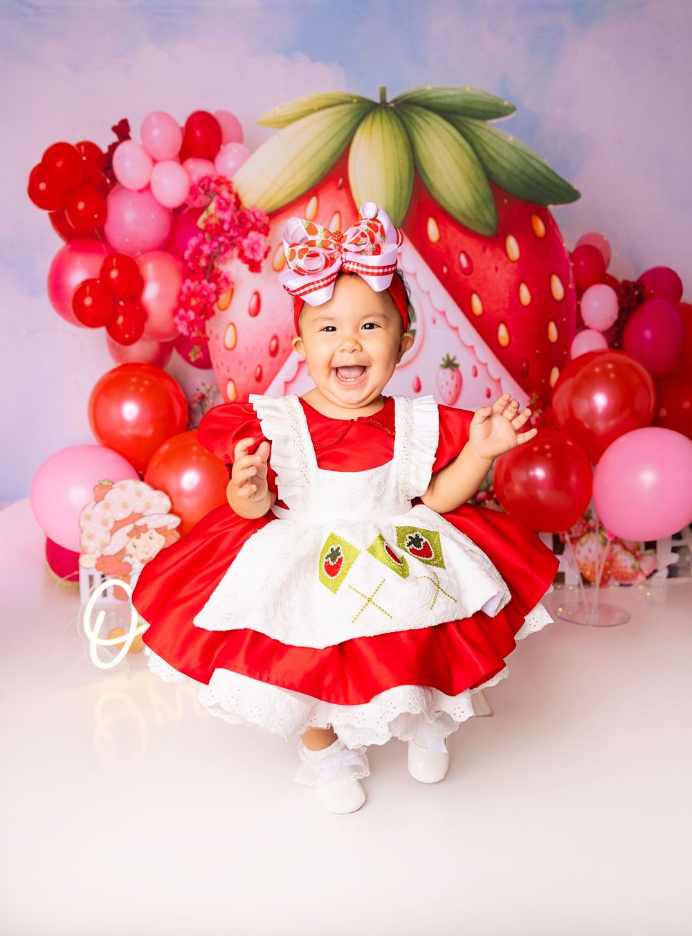 Baby girl in red Strawberry Shortcake dress smiles at first birthday cake smash with strawberry balloon backdrop.