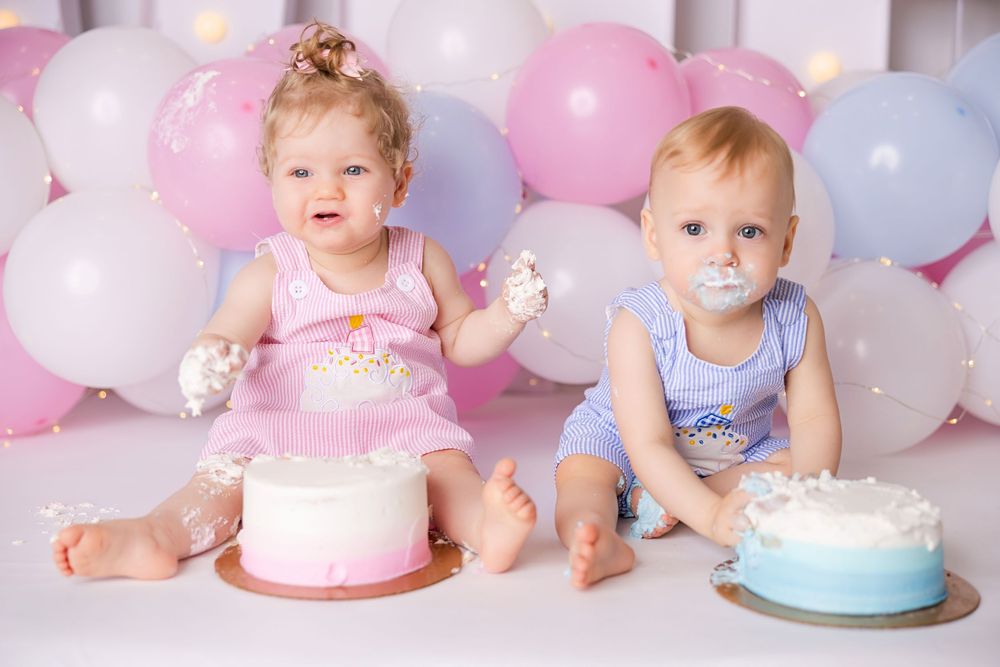 Two babies in pink and blue outfits sit with birthday cakes surrounded by pastel balloons at a celebration.
