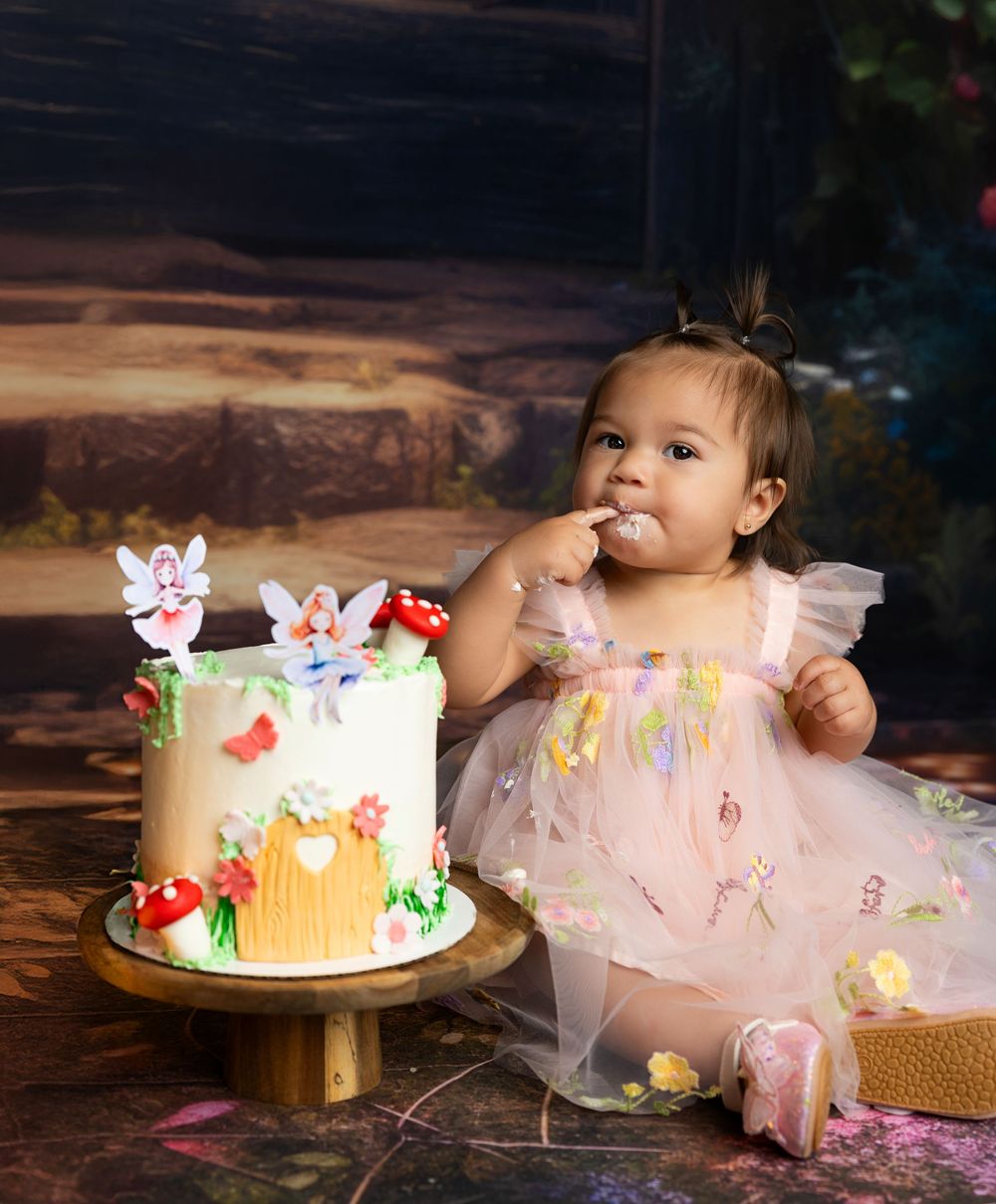 Little one in pink tulle dress enjoys their first birthday celebration with a decorated butterfly cake on wooden table.