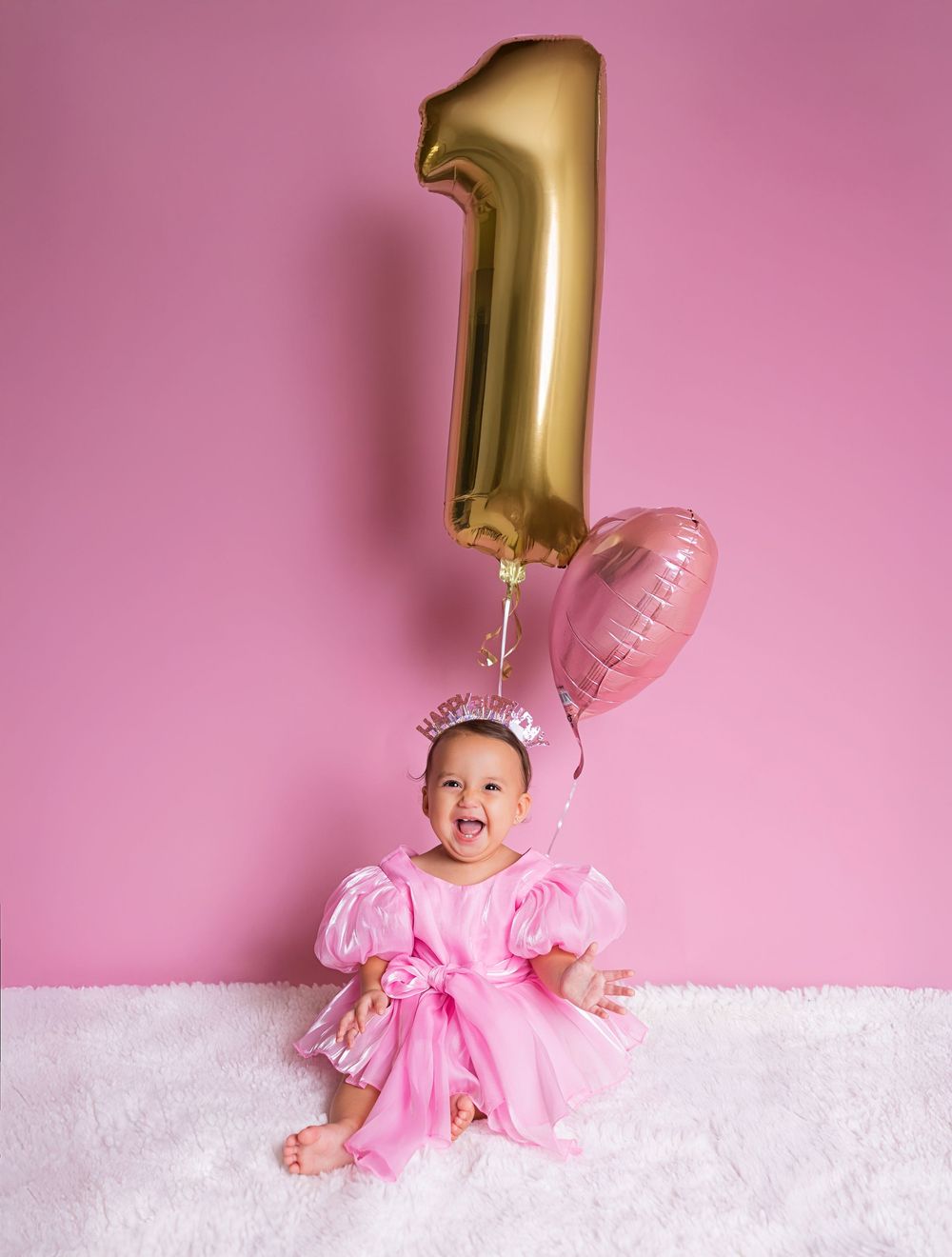 Baby girl in pink princess dress celebrates first birthday with gold number one and pink balloons on white fur rug.